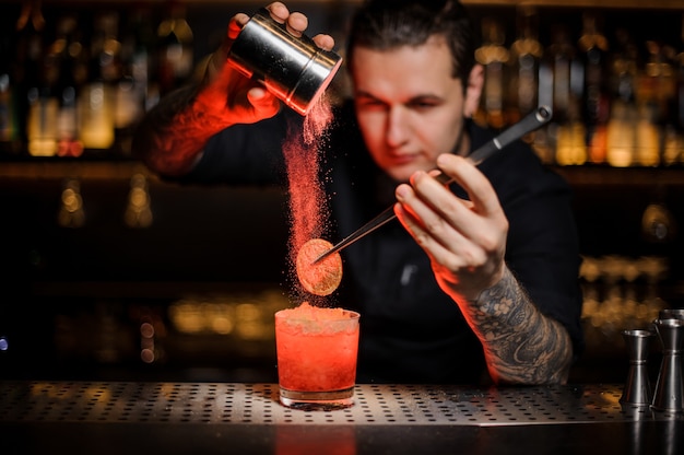 Professionele barman die aan een alcoholische cocktail in het glas een gedroogde sinaasappel toevoegt met een pincet en aromatisch poeder in het rode licht op de toog.