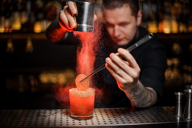 Professionele barman die aan een alcoholische cocktail in het glas een gedroogde sinaasappel toevoegt met een pincet en aromatisch poeder in het rode licht op de toog.