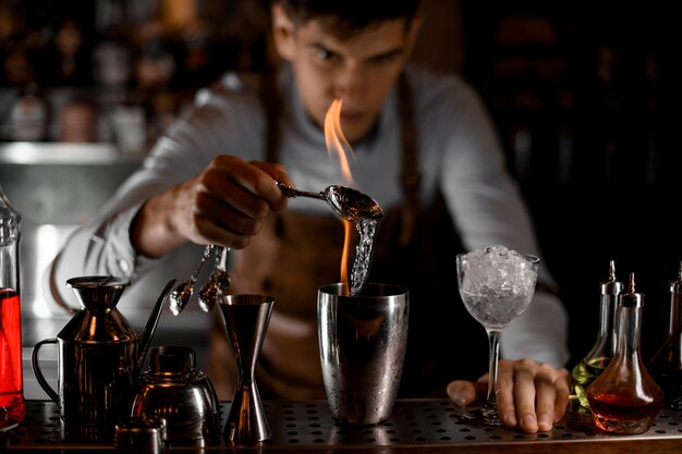 Foto professionele barman gieten een essentie van de lepel in de vlam naar de stalen shaker