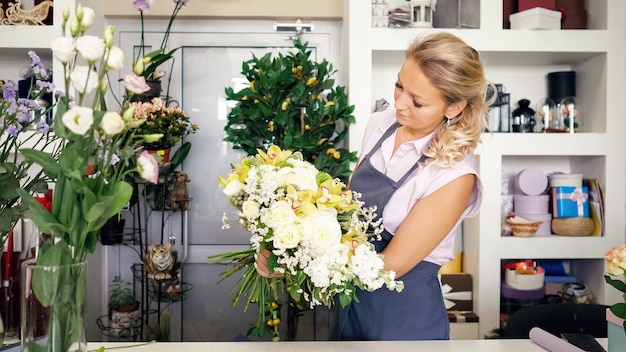 Professionele bloemistvrouw maakt een prachtig bloemboeket met rozen, bladeren en gipskruid. Blonde vrouw van middelbare leeftijd in uniform werkt in bloemenwinkel. Ze maakt een heleboel tedere kleuren roze en geel.