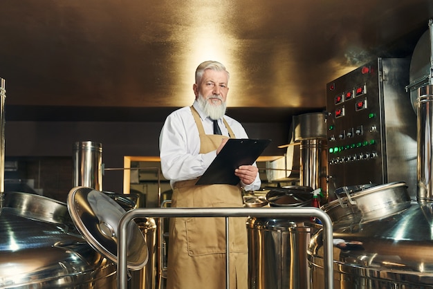 Professionele brouwerijmedewerker die het proces van het brouwen van bier controleert. Knap, bejaarde, bebaarde arbeider die in de map van de schortholding, camera bekijkt.