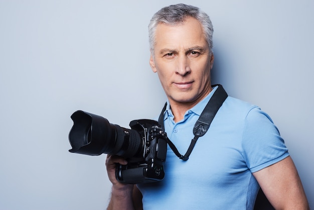 Professionele fotograaf. Portret van zelfverzekerde volwassen man in T-shirt die camera vasthoudt terwijl hij tegen een grijze achtergrond staat