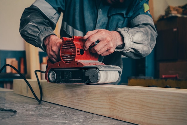 Professionele houtbewerker maalt houten staaf op werkbank in timmerwerkplaats met gereedschap Middelbare leeftijd werknemer in overall verwerkt hout met elektrische slijper Echte workflow