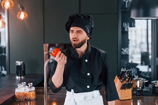 Professionele jonge chef-kok in uniform staat bij de tafel en poseert voor een camera in de keuken