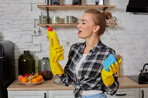 Professionele jonge vrouw die rubberen beschermende gele handschoenen draagt met flessenreinigers in de keuken.