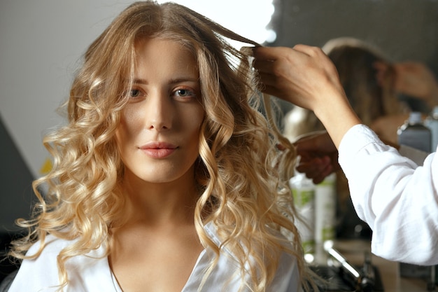 Professionele kapper coiffeur krullen met ijzer maken tot een modieuze blonde vrouw in schoonheidssalon. close-up shot met schaduw