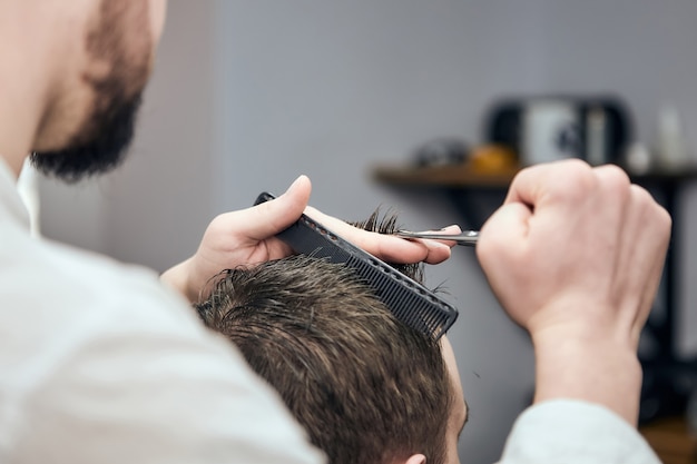 Professionele kapper die kapsel maakt voor jonge man met schaar en kam bij kapperszaak