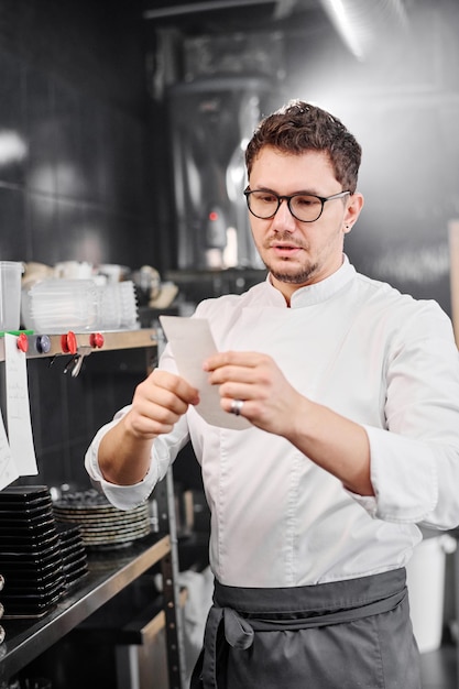 Professionele kok in schort die papier in zijn handen houdt en de volgorde van de gast onderzoekt voordat hij kookt