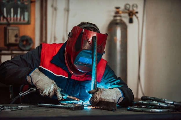 Professionele lasser in beschermend uniform en maskerlassen van metalen buizen op de industriële tafel met andere arbeiders erachter in de industriële werkplaats