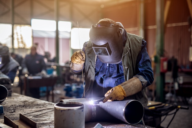 Professionele lasser in beschermende uniform en masker lassen van metalen pijp op de industriële tafel met andere werknemers achter in de industriële werkplaats.