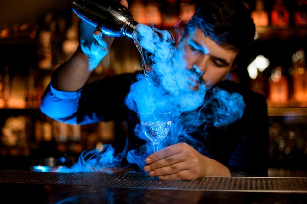 Professionele mannelijke barman die een rook in het cocktailglas van de schudbeker gieten onder het blauwe licht