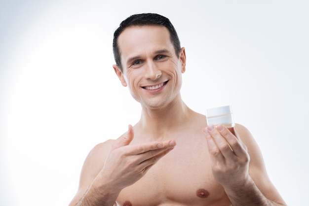 Professionele mannelijke cosmetica. Vrolijke, aangename jongeman die lacht en naar je kijkt terwijl hij een gezichtscrème presenteert