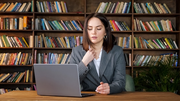 Professionele secretaresse in grijze kostuumtypes op laptop zittend onder sterke ventilatorwind tegen verschillende gekleurde boeken op rekken in kantoor