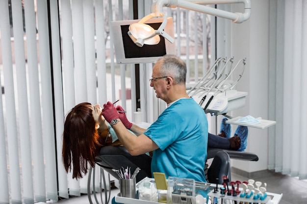 Professionele senior man arts tandarts doet een tandheelkundige behandeling op een jonge vrouwelijke patiënt in moderne tandheelkundige kliniek
