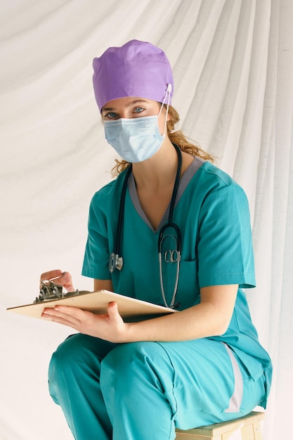 Professionele vrouwelijke arts in scrubs en medisch masker en met een stethoscoop zittend op een houten kruk en kijkend naar de camera tijdens het controleren van aantekeningen op klembord op beige achtergrond