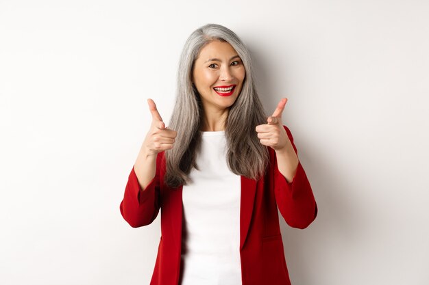 Professionele vrouwelijke werkgever in trendy rode blazer en make-up, met de vingers naar de camera wijzend en glimlachen, iets prijzend, heb je nodig, staande op een witte achtergrond