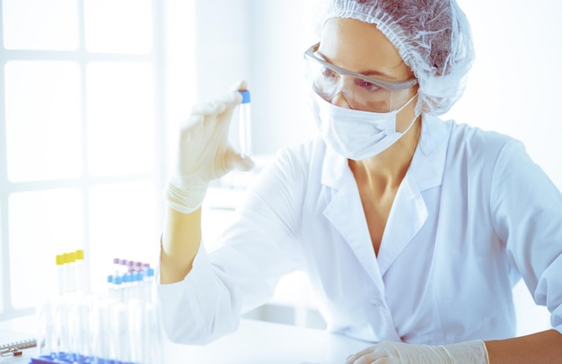 Professionele vrouwelijke wetenschapper in beschermende brillen onderzoeken buis met reagentia in zonnig laboratorium afgezwakt in blauw Geneeskunde en wetenschap onderzoek