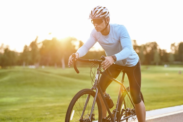 Professionele wielrenner in sportkleding en beschermende helm die bij zonsondergang op de weg staat