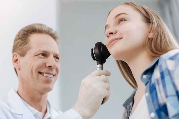 Photo proficient aged delighted doctor enjoying responsibilities in the clinic and sitting in the cabinet while examining patient skin with dermatoscope