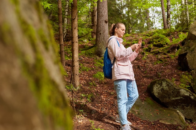 Profiel die van Europese reiziger roze jasje, blauwe rugzak, jeans en tennisschoenen dragen, smartphone in handen houden, foto nemen, die verbinding zoeken, kamperen tijdens vakantie. Wandelen concept.