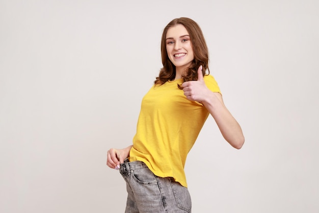 Profiel portret van mooie gelukkige tiener meisje wilskracht tonen slanke taille in grote broek en duimen opdagen succesvol gewichtsverlies indoor studio shot geïsoleerd op grijze achtergrond