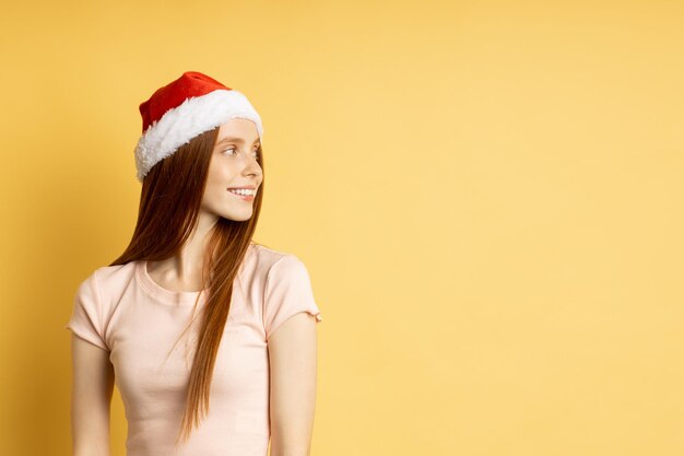 Profiel shot van serieuze peinzende aantrekkelijke roodharige vrouw met sproeten, gekleed in t-shirt, kerstmuts staande met gekruiste handen, op zoek naar kant geïsoleerde gele achtergrond met kopie ruimte.