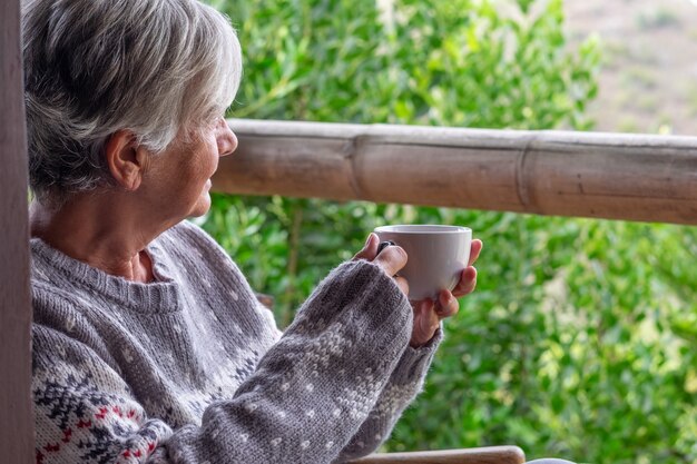 Profiel van een grijsharige oudere vrouw in een wintertrui zittend op het houten balkon kijkend naar het bos met een kopje koffie.
