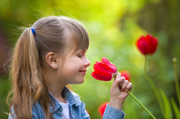 Profiel van leuk mooi glimlachend kindmeisje met grijze ogen en lang haar die heldere rode tulpenbloem ruiken op de vage zonnige achtergrond van de zomer groene bokeh.