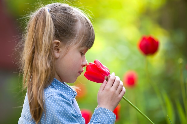 Profiel van leuk mooi glimlachend kindmeisje met grijze ogen en lang haar die heldere rode tulpenbloem ruiken op vage zonnige de zomer groene bokeh