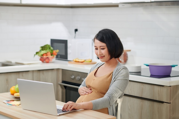 Profiel zijaanzicht aantrekkelijke en progressieve jonge mama zittend in een stoel achter tafel in lichte flat en met behulp van pc-laptop