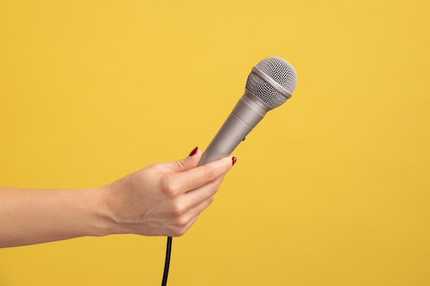 Profiel zijaanzicht close-up van menselijke hand met zilveren microfoon voor zingen of rapporteren Indoor studio shot geïsoleerd op gele achtergrond