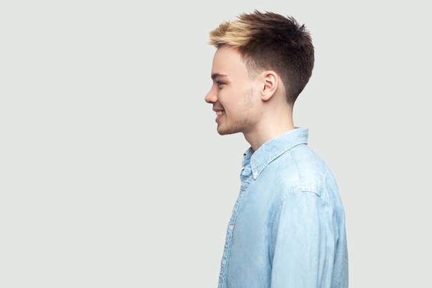 Profiel zijaanzicht portret van een gelukkige knappe jongeman in een lichtblauw shirt dat staat met een brede glimlach en vooruitkijkt met geluk. indoor studio opname op grijze achtergrond kopie ruimte.