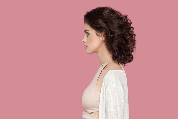 Profiel zijaanzicht portret van rustige mooie brunette jonge vrouw met krullend kapsel in casual stijl permanent met serieus gezicht en kijken. indoor studio opname geïsoleerd op roze achtergrond.