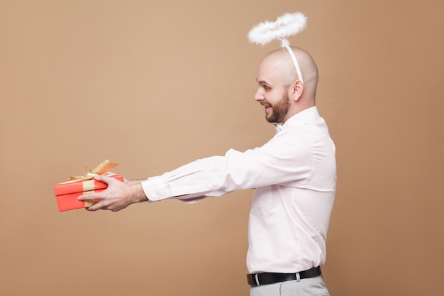 Profiel zijaanzicht van een gelukkige knappe kale bebaarde engel van middelbare leeftijd in shirt en witte halo op het hoofd die staat en een rood cadeau geeft met een brede glimlach. studio opname, geïsoleerd op lichtbruine achtergrond.