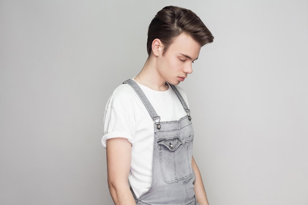 Profiel zijaanzicht van trieste jonge brunette man in casual stijl met t-shirt en denim overalls staan en houden het hoofd naar beneden met gesloten ogen en verdriet. indoor studio-opname, geïsoleerd op een grijze achtergrond