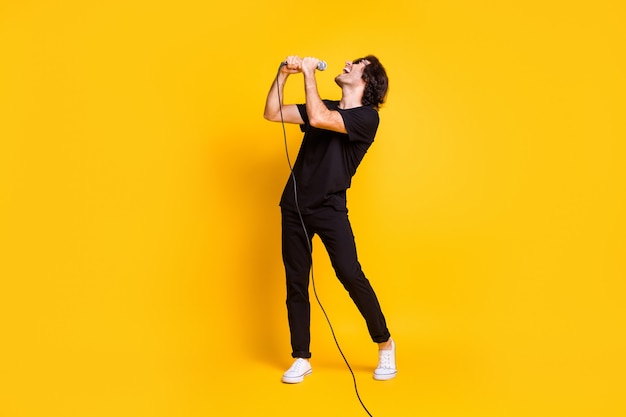 Profielfoto van de volledige lengte van een jonge man die de microfoon vasthoudt en open mond zingt, een zwarte t-shirtbroek draagt, een witte sneakers, een bril, een geïsoleerde gele kleurachtergrond