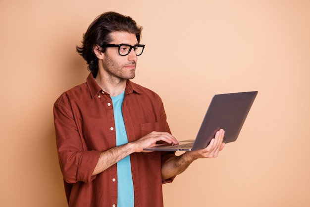 Foto profielfoto van een serieuze kerel die een computerscherm vasthoudt, een bril draagt een bruin t-shirt geïsoleerd beige kleur achtergrond