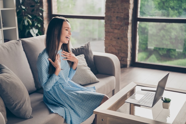 Profielfoto van mooie vrolijke dame praten skype notitieboekje online videogesprek datum vriendje vergadering doorbrengen quarantaine thuis blijven zitten bank dragen chique gestippelde jurk woonkamer binnenshuis