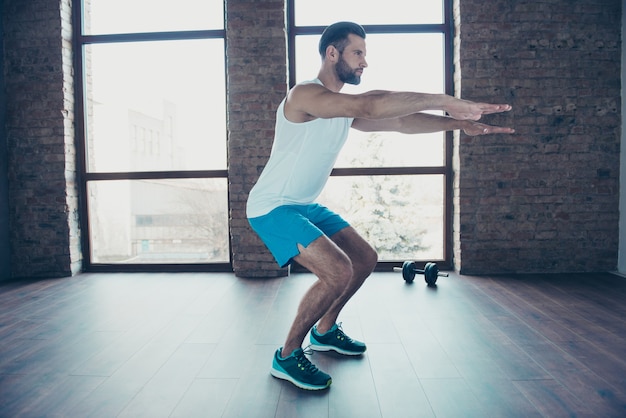 Profielfoto van volledige lengte van macho man die statische squats doet, vetverbrandingsproces, sportkleding, tank-top, korte broek, sneakers, opleiding, huis, thuis, dichtbij, grote ramen, binnen