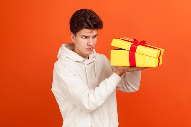 Profielportret van twijfelachtige tiener met modieus kapsel in casual trui die in geschenkdoos kijkt, glurend. Indoor studio-opname geïsoleerd op oranje achtergrond