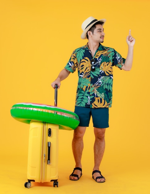 Profielschot, jonge aziatische man in kleurrijk hawaiiaans overhemd geeft wijsvinger op en trekt gele koffer. full body studio portret op gele achtergrond. gelukkig zomer vakantie reizen concept.