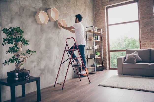 Profielweergave op volledige grootte van een man van gemengd ras die wandplank installeert staande trapladder reparatie van nieuwe platte handgemaakte werkende hangende laatste plank duurzaamheid plat binnenshuis controleren