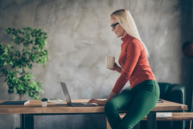 Profielzijde foto van coll slimme deskundige vrouw marketeer zitten bureau houden koffiekopje werken op computer opstartinformatie lezen statistieken op zolder kantoor werkplek dragen rode coltrui