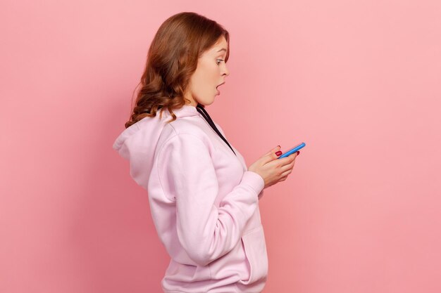 Profile of amazed curly haired teenage girl looking at\
cellphone with surprised expression reading shocking news using\
mobile phone indoor studio shot isolated on pink background