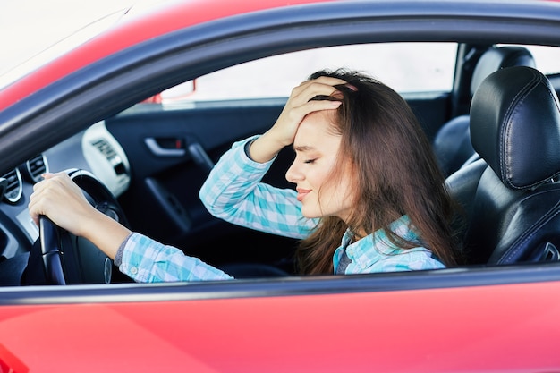 profile of annoyed woman driving red car, stress while driving. tensed woman leaning on hand with closed eyes, traffic jams. head and shoulders of brunette woman inside car