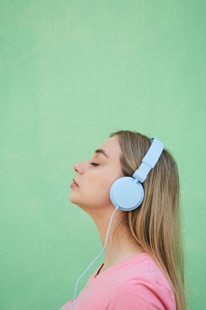 Profile of a blonde woman listening to music in blue headphones against a green wall