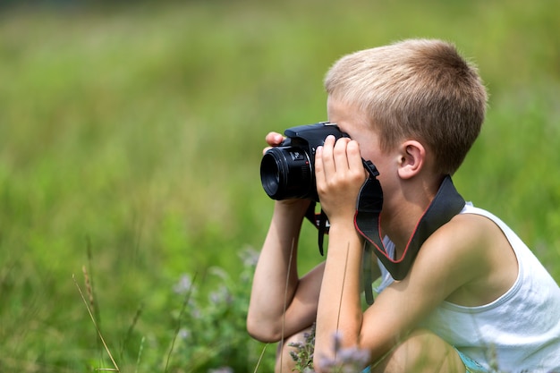 Profile close-up portrait of young blond cute handsome child boy with camera taking pictures outdoors on bright sunny spring or summer day on blurred light green grassy