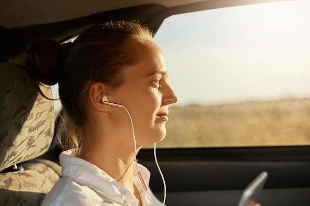 Profile closeup portrait of beautiful business woman is using a smart phone and earphone for listening music smiling while sitting on back seat in the car looking at window at sunset