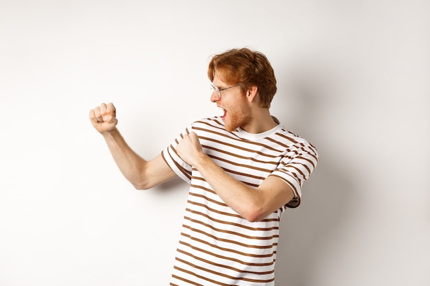 Profile of funny young man with red hair and beard, raising fists for fight, shadow boxing and shouting, standing over white background