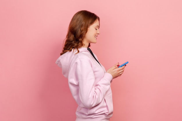 Profile of happy curly haired teenage female in casual hoodie sincerely smiling while using smartphone messaging entertainment app Indoor studio shot isolated on pink background